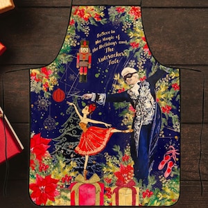 Puede incluir: Un delantal de Navidad azul y dorado con temática de Cascanueces. Presenta una bailarina bailando en un árbol de Navidad, un soldado Cascanueces y el texto "Cree en la magia de las fiestas y el cuento del Cascanueces".