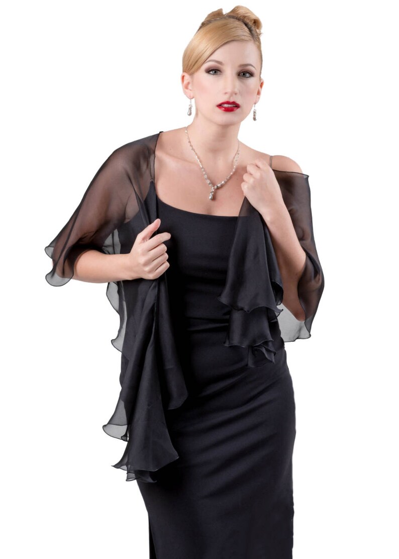 May include: A woman wearing a black dress with a sheer black shawl. The shawl has ruffled edges and is draped over her shoulders. She is wearing a silver necklace and earrings.
