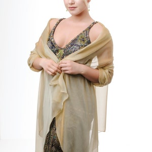 May include: A woman wearing a gold sheer shawl over a patterned dress. The shawl is draped over her shoulders and tied at the front. She is looking at the camera.