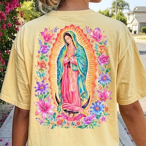 May include: A pale yellow short-sleeve t-shirt featuring a vibrant graphic on the back of the Virgin of Guadalupe surrounded by colorful flowers. The shirt is paired with a bright pink baseball cap and a matching pink shoulder bag.