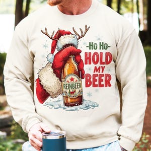 Puede incluir: Sudadera beige con un diseño navideño. El diseño presenta a Papá Noel sosteniendo una botella de cerveza con el texto "-Ho Ho- HOLD MY BEER". La botella de cerveza está etiquetada como "Reinbeer".