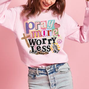 May include: A light pink sweatshirt with the words "pray more, worry less" in a colorful, collage-style design. The graphic includes a wooden cross, a heart, praying hands, and a peace symbol. The sweatshirt is paired with light-wash denim jeans.