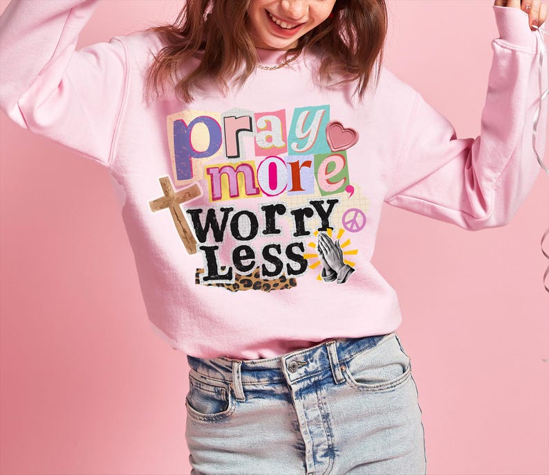 May include: A light pink sweatshirt with the words "pray more, worry less" in a colorful, collage-style design. The graphic includes a wooden cross, a heart, praying hands, and a peace symbol. The sweatshirt is paired with light-wash denim jeans.