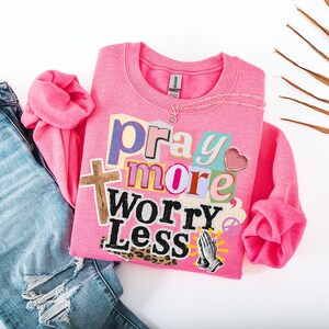 May include: A pink sweatshirt with the words "Pray More Worry Less" in a collage style, paired with blue denim jeans. The sweatshirt features a cross, heart, and praying hands graphic. A silver necklace is on the sweatshirt.