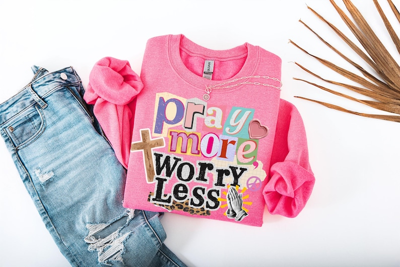 May include: A pink sweatshirt with the words "Pray More Worry Less" in a collage style, paired with blue denim jeans. The sweatshirt features a cross, heart, and praying hands graphic. A silver necklace is on the sweatshirt.