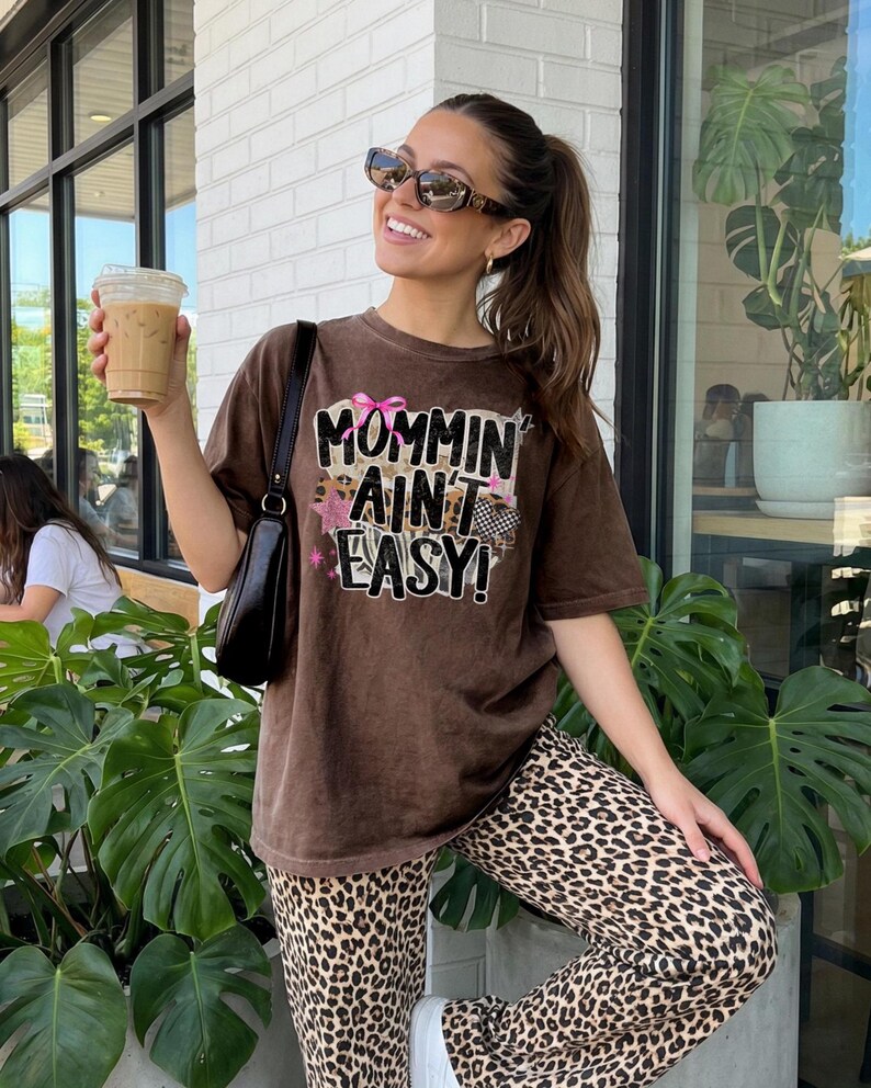 May include: Brown t-shirt with the text "Mommin Ain't Easy!" paired with leopard print pants. The image shows a woman wearing sunglasses and holding a beverage. The shirt has a graphic design with pink and black accents.
