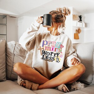 May include: Beige sweatshirt with the text "Pray More Worry Less" in colorful letters. The sweatshirt features a cross, a peace sign, and a praying hands graphic. A black mug is held up to the face.