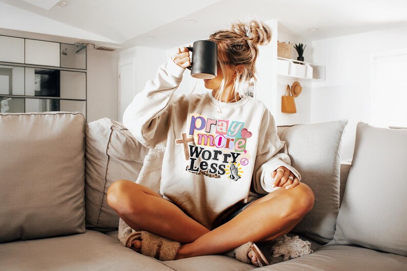 May include: Beige sweatshirt with the text "Pray More Worry Less" in colorful letters. The sweatshirt features a cross, a peace sign, and a praying hands graphic. A black mug is held up to the face.