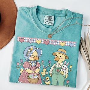 May include: A turquoise Comfort Colors t-shirt featuring an Easter-themed graphic. The design includes two duck characters, one holding a basket of eggs and the other holding a bunny. A necklace with a pendant is draped across the shirt.