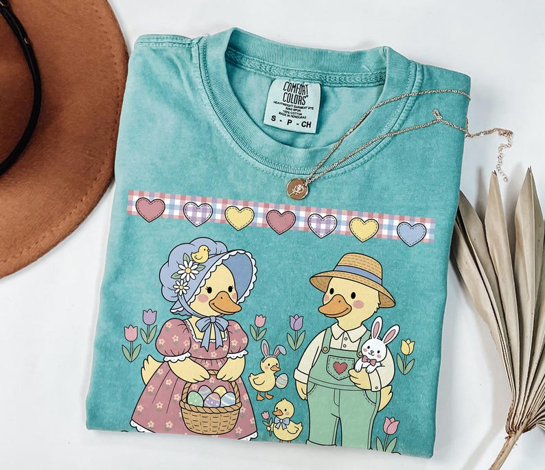 May include: A turquoise Comfort Colors t-shirt featuring an Easter-themed graphic. The design includes two duck characters, one holding a basket of eggs and the other holding a bunny. A necklace with a pendant is draped across the shirt.