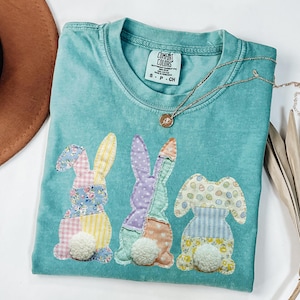 Op de afbeelding: Een turquoise Comfort Colors t-shirt met drie patchwork konijn ontwerpen met bloemen-, stippen- en streepjespatronen. Elk konijn heeft een pluizige witte pomponstaart. Een gouden ketting met een hanger is over het shirt gedrapeerd.
