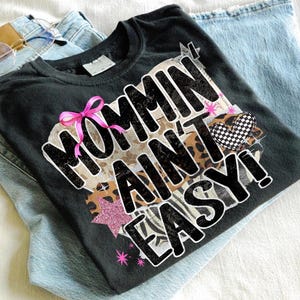 May include: Black t-shirt with the phrase "Mommin Ain't Easy!" in a bold, stylized font. The design includes a pink bow, a checkered heart, and a star. The shirt is folded on top of a pair of light blue jeans.