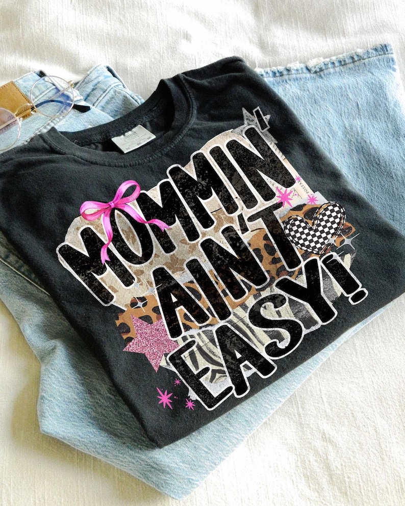 May include: Black t-shirt with the phrase "Mommin Ain't Easy!" in a bold, stylized font. The design includes a pink bow, a checkered heart, and a star. The shirt is folded on top of a pair of light blue jeans.