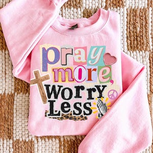 May include: A light pink sweatshirt with the words "pray more, worry less" in a collage style. The design includes a wooden cross, a heart, praying hands, and a peace sign.