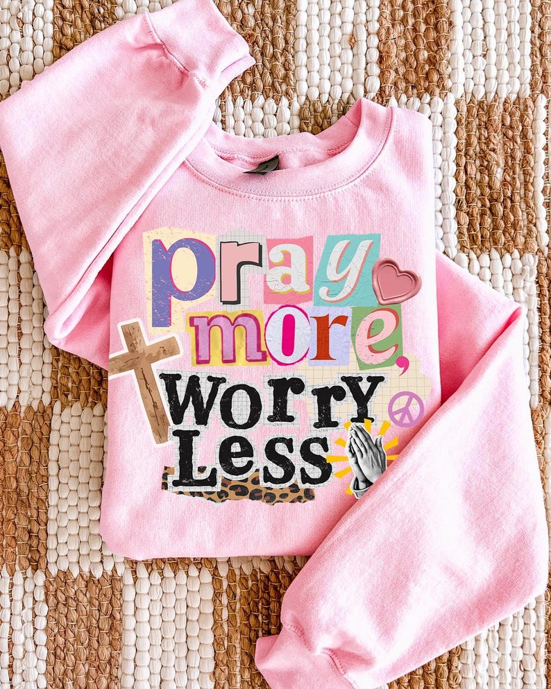 May include: A light pink sweatshirt with the words "pray more, worry less" in a collage style. The design includes a wooden cross, a heart, praying hands, and a peace sign.
