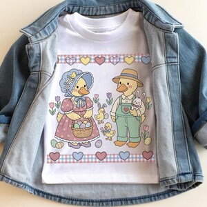 May include: White t-shirt featuring an Easter-themed graphic. The design includes two cartoon ducks, one wearing a bonnet and holding a basket of eggs, and the other wearing overalls and a straw hat, holding a bunny. The shirt is displayed on a denim jacket.