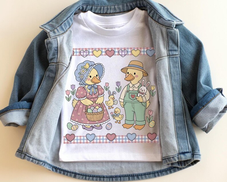 May include: White t-shirt featuring an Easter-themed graphic. The design includes two cartoon ducks, one wearing a bonnet and holding a basket of eggs, and the other wearing overalls and a straw hat, holding a bunny. The shirt is displayed on a denim jacket.