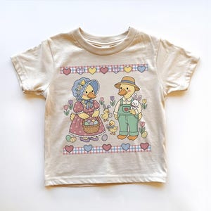 May include: Beige toddler t-shirt featuring a charming Easter-themed graphic. The design includes two ducks dressed in vintage attire, surrounded by Easter eggs, flowers, and a bunny. The shirt has a crew neck and short sleeves.