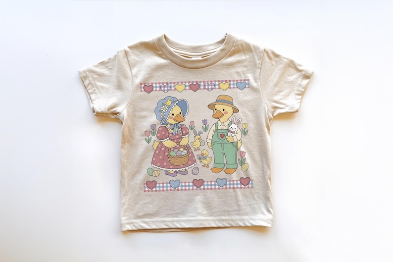 May include: Beige toddler t-shirt featuring a charming Easter-themed graphic. The design includes two ducks dressed in vintage attire, surrounded by Easter eggs, flowers, and a bunny. The shirt has a crew neck and short sleeves.