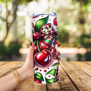 Puede incluir: Un vaso blanco con un dise&ntilde;o de cerezas. El dise&ntilde;o incluye cerezas rojas, hojas verdes, un lazo con estampado de leopardo y peque&ntilde;os corazones. El vaso tiene una tapa plateada y una pajita blanca.