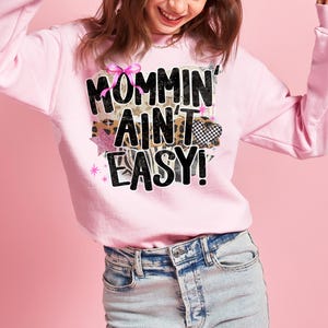 May include: A light pink sweatshirt with the phrase "Mommin' Ain't Easy!" in a mix of fonts and patterns. The design includes a pink bow, leopard print, and a checkered heart. The sweatshirt is paired with light-wash denim jeans.