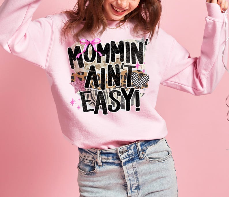 May include: A light pink sweatshirt with the phrase "Mommin' Ain't Easy!" in a mix of fonts and patterns. The design includes a pink bow, leopard print, and a checkered heart. The sweatshirt is paired with light-wash denim jeans.