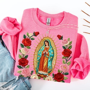 Puede incluir: Sudadera rosa de cuello redondo con una imagen bordada de la Virgen María, rodeada de rosas rojas y rosas. Un collar plateado descansa sobre la camisa, junto a unos vaqueros azules.