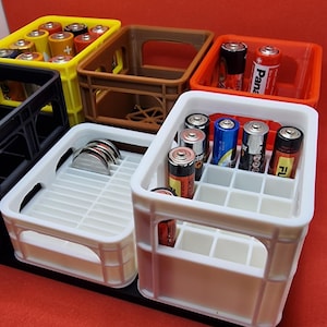 Puede incluir: Cuatro cajas de plástico blanco con diferentes compartimentos para organizar artículos. La caja superior tiene una variedad de baterías, incluyendo Duracell, Energizer y Panasonic. Las otras cajas están vacías.