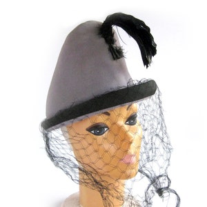 May include: A gray felt hat with a black band and a black feather. The hat is on a mannequin head and has a black veil.