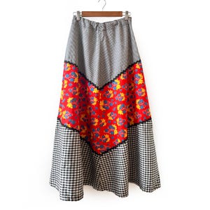 以下が含まれることがあります： パッチワークデザインのロングスカート。上部と下部は白黒のチェック柄で、中央部は赤い花柄です。黒いトリムがセクションを区切っています。スカートは木製のハンガーから吊り下げられています。