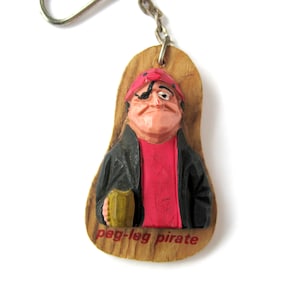 May include: Wooden keychain featuring a cartoon pirate with a red hat, eyepatch, and a black jacket holding a mug. The keychain is labeled "peg-leg pirate".