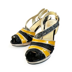 May include: A pair of high-heeled sandals with a platform sole. The sandals feature a black, yellow, and silver glitter design. The straps are black and yellow patent leather, with silver glitter accents. The heels are approximately 4 inches high.