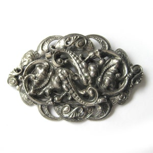 Puede incluir: Un broche de filigrana de tono plateado con un diseño detallado e intrincado. El broche tiene forma ovalada y presenta un patrón floral en espiral.