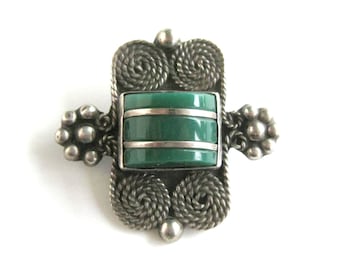 Puede incluir: Un broche de plata con una piedra verde engastada en un marco cuadrado. El marco está decorado con intrincados trabajos de alambre de plata.