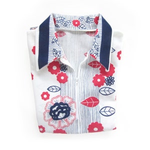 May include: A white collared shirt with a blue trim and a zipper closure. The shirt has a floral pattern with red and blue flowers and leaves.