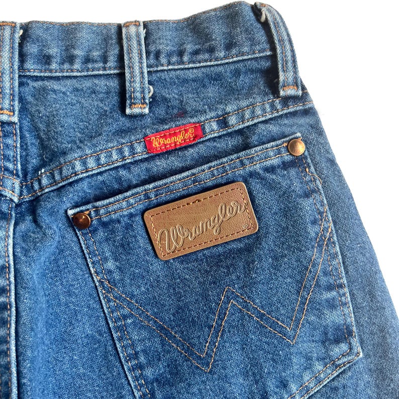 May include: Close-up of the back of blue denim jeans. Features a brown leather patch with the Wrangler logo, a red Wrangler tag, and a back pocket with a stitched W design. The jeans have a classic, vintage look.