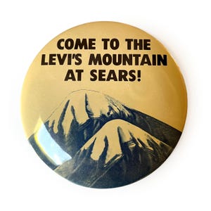 May include: A round, gold-toned button with the text "COME TO THE LEVI'S MOUNTAIN AT SEARS!" in bold, dark brown letters. Below the text is a graphic of a mountain range in shades of blue and gray.