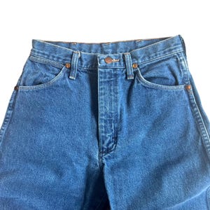 May include: Close-up of blue denim jeans. The jeans have a classic five-pocket design, a button and zipper closure, and copper-colored rivets. The denim has a medium wash and a slightly worn appearance.