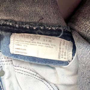 May include: Close-up of a Wrangler jeans label. The label is white with black text, including "Wrangler", size information, and care instructions. The jeans are blue denim, with visible stitching and a button.