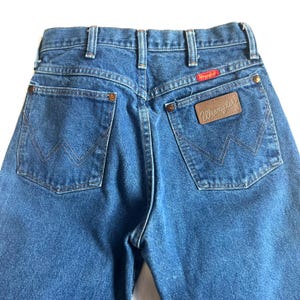 May include: Close-up of the back of blue denim jeans. The jeans have two back pockets with a stitched design, a brown leather patch with the brand name "Wrangler", and a red tag with the brand name. The jeans are a classic style.