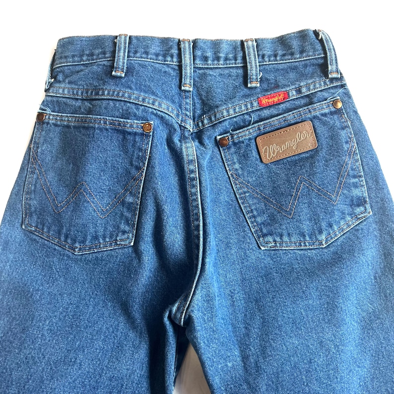 May include: Close-up of the back of blue denim jeans. The jeans have two back pockets with a stitched design, a brown leather patch with the brand name "Wrangler", and a red tag with the brand name. The jeans are a classic style.