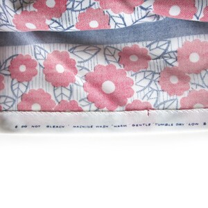 May include: Close-up of a white fabric with a pink and blue floral pattern. The fabric has a care label that reads "Do not bleach, machine wash warm, gentle tumble dry low."