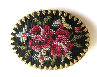 May include: An oval-shaped brooch with a gold-colored metal frame and a black fabric center. The fabric is embroidered with a floral design in shades of pink, red, and blue.
