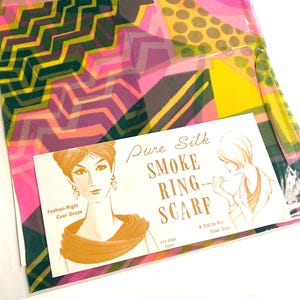 May include: A colorful silk scarf with a geometric pattern in pink, yellow, green, and purple. The scarf is packaged with a card that reads "Pure Silk Smoke Ring Scarf" and features illustrations of women.