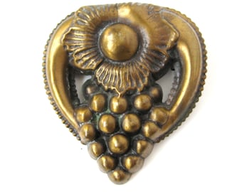 May include: A gold-toned metal brooch in the shape of a grape cluster with a stylized flower design. The brooch is detailed with a textured surface and a raised design.