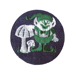 May include: A green and white embroidered patch of a smiling elf holding a white mushroom on a dark blue denim background.