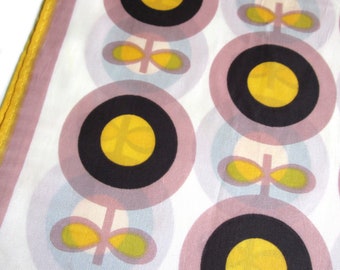 May include: A white fabric scarf with a repeating pattern of pink circles containing black circles with yellow centers and a stylized green leaf design.