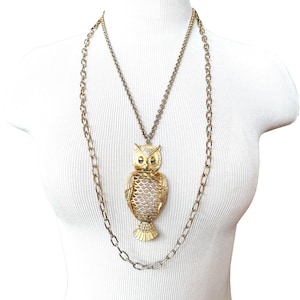 May include: A long gold-tone necklace featuring a large owl pendant. The owl has textured wings with a scale-like pattern, prominent ears, and circular eyes. The pendant hangs from a delicate chain, which is layered over a longer, thicker oval link chain.