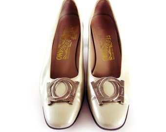 May include: A pair of off-white leather pumps with a silver buckle featuring the Salvatore Ferragamo logo. The shoes have a low heel.