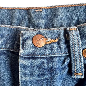 May include: Close-up of blue denim jeans with a copper-colored button and a buttonhole. The button has a decorative design. The stitching is in a contrasting orange-brown color.